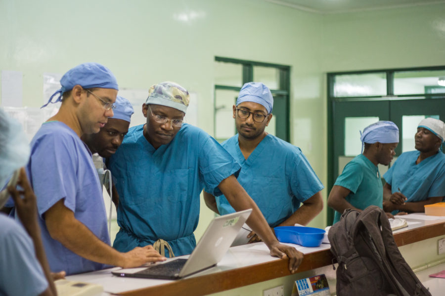 Doctors in blue scrubs stand around a laptop.
