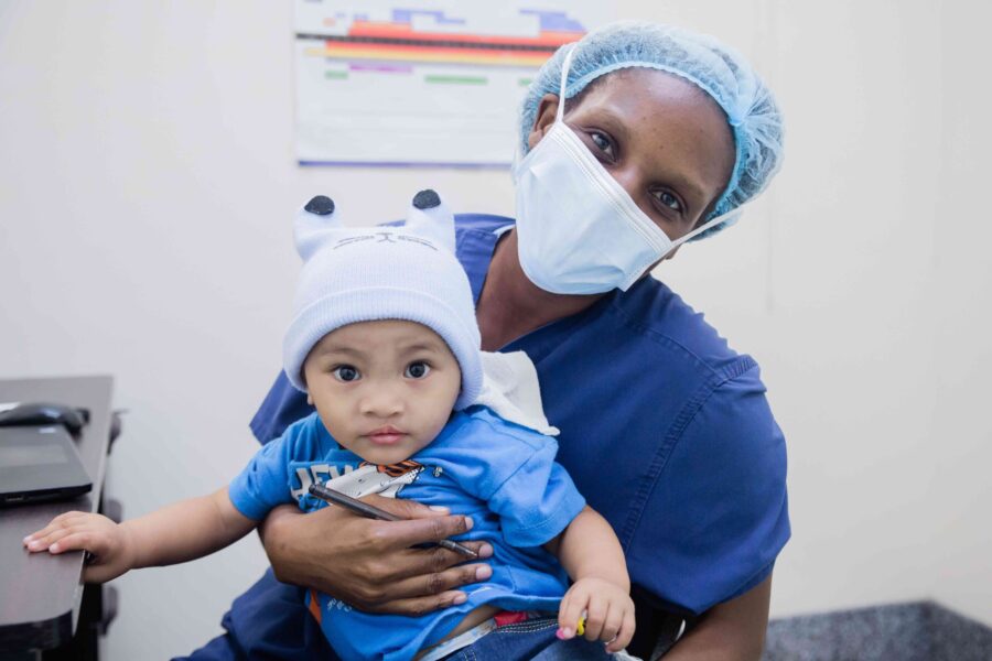 A female healthcare professional wearing a surgical mask and hairnet holding a child.